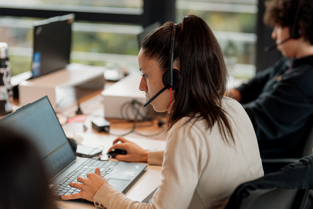 Una mujer con auriculares y micrófono trabaja en su laptop en una oficina. Está concentrada en la pantalla, con su mano en el ratón y la otra en el teclado.
