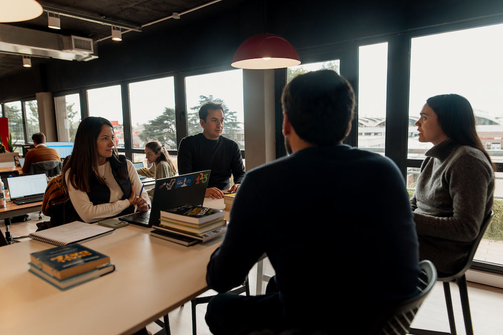 Un grupo de cuatro personas conversa alrededor de una mesa en una oficina abierta. Hay laptops y libros sobre la mesa, y grandes ventanas en el fondo.